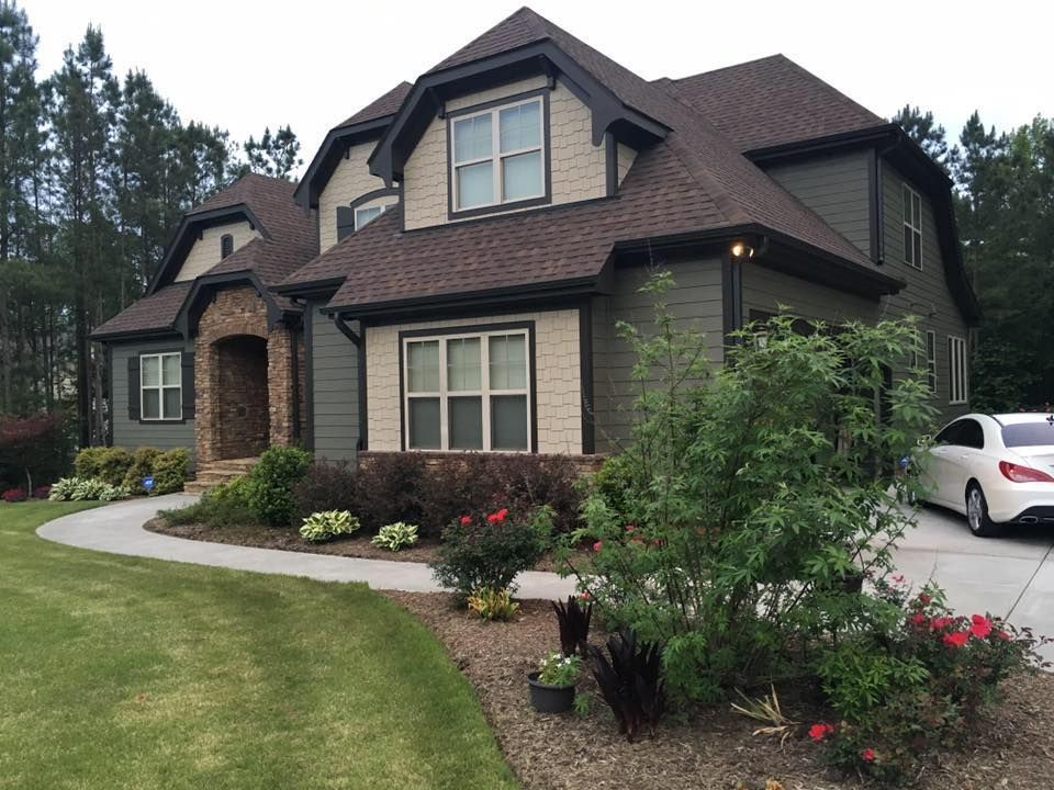 Large two-story suburban house with stone facade, brown roof, manicured lawn, and shrubs, with a white car in driveway