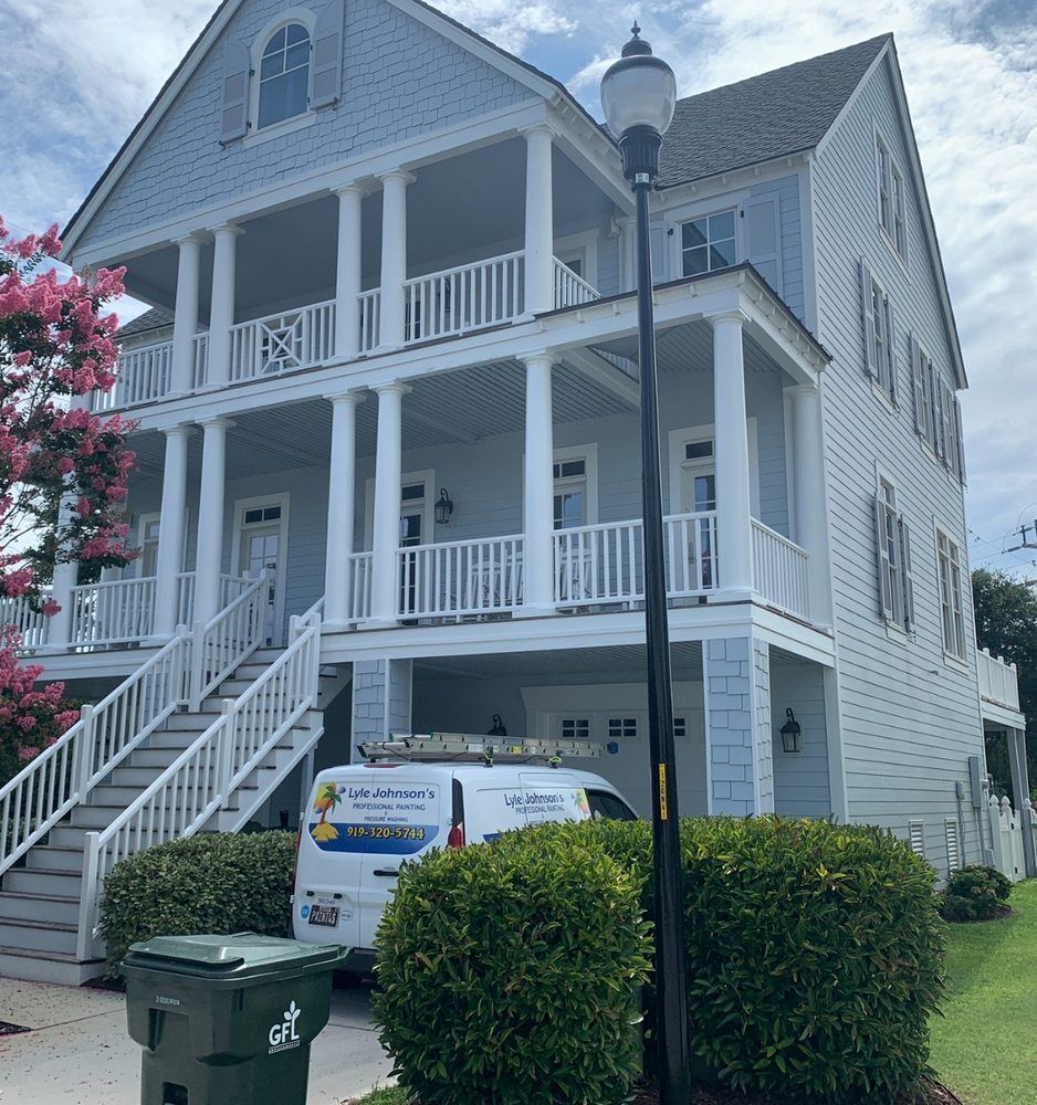 White three-story house with porches, stairs, a white van, and trimmed hedges on a sunny day
