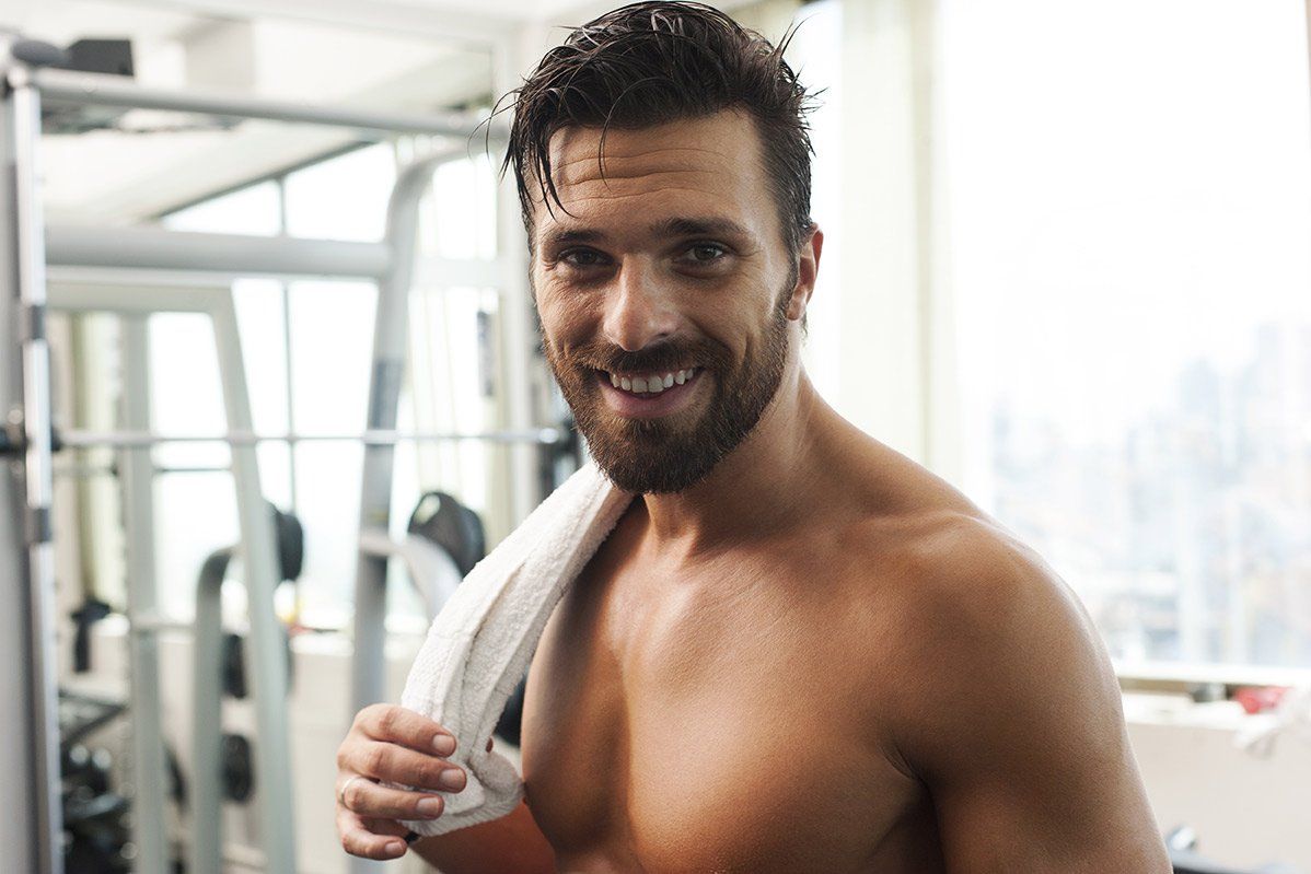A shirtless man with a towel around his neck is smiling in a gym.