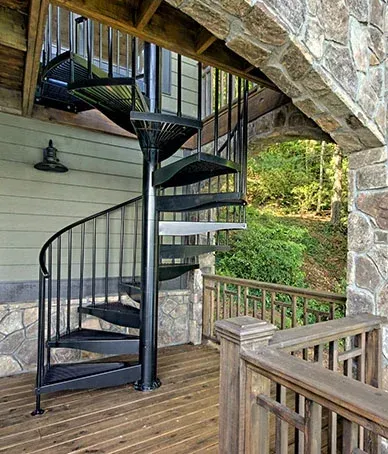 Black spiral staircase on a wooden deck, leading to a covered porch with stone columns and greenery in the background.