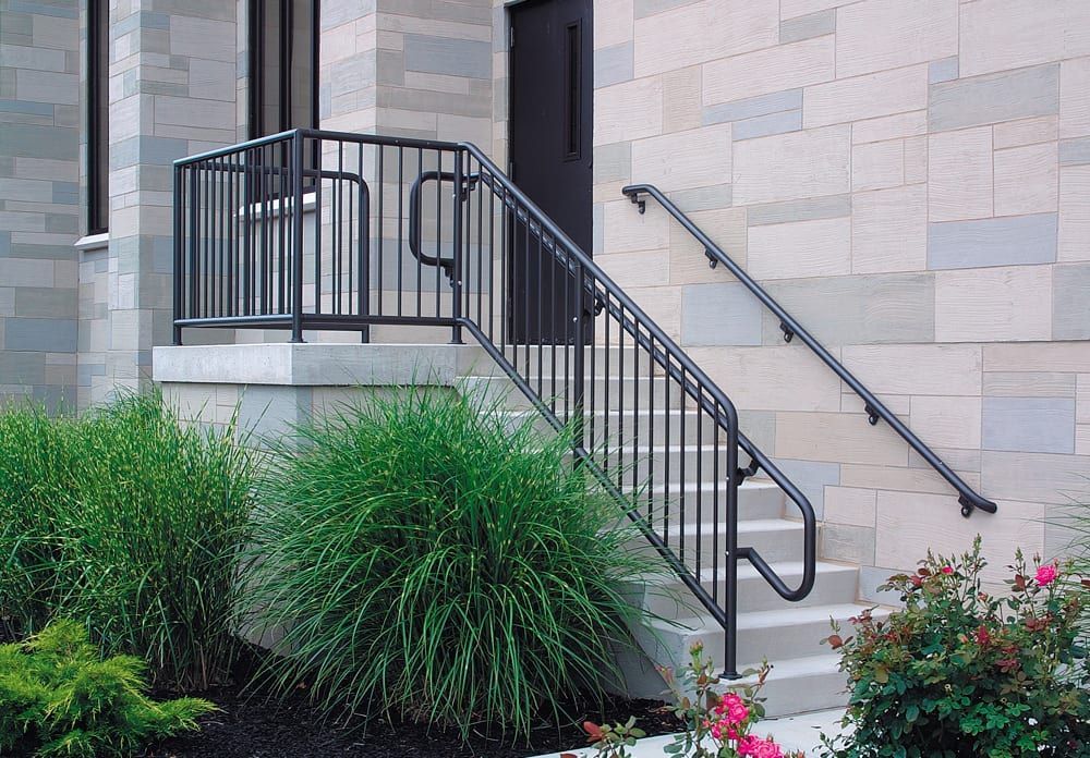 Exterior steps with black metal handrails and a landing. Light stone building with shrubs.