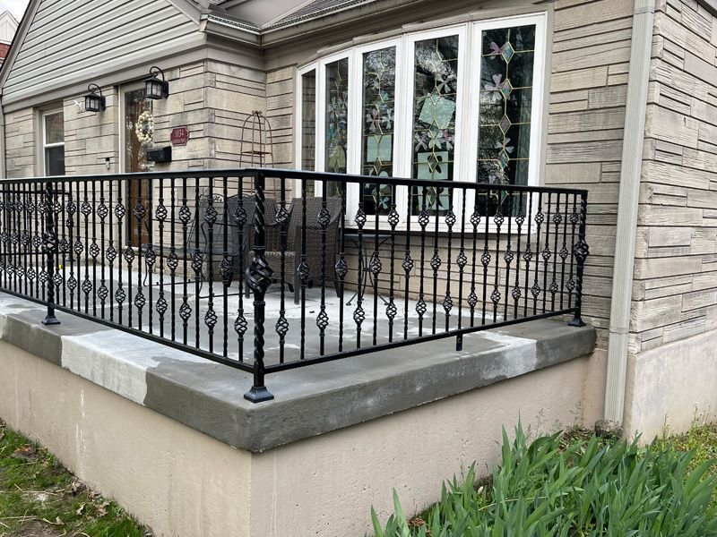 Black metal railing on a stone porch, in front of a house with a bay window.