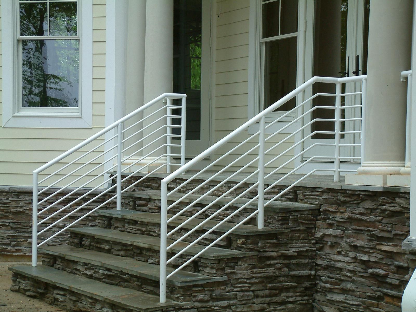 White metal railing on stone steps leading to a house with white pillars and windows.