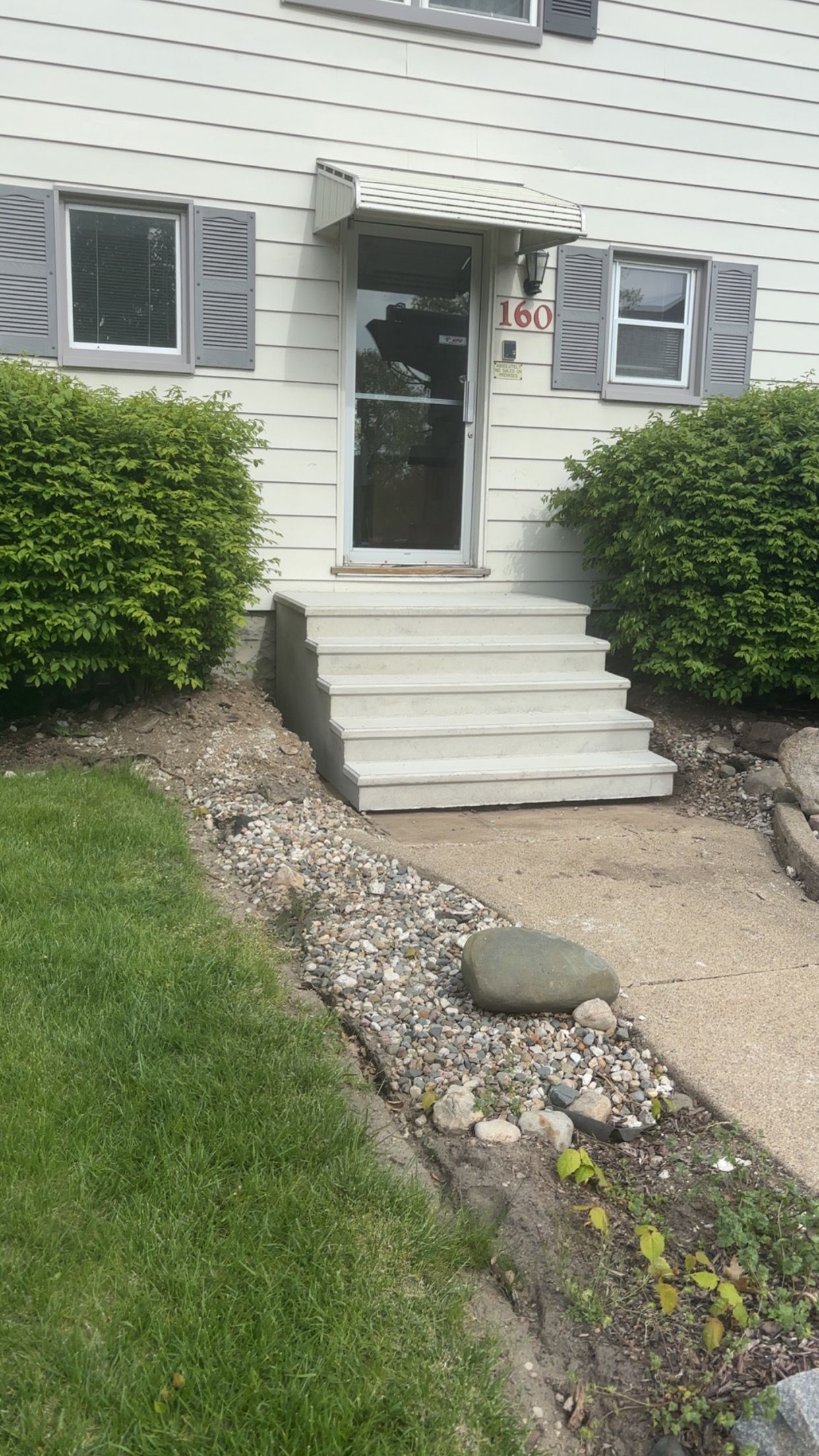 White house with gray shutters, concrete steps, and a glass door. Landscaping includes bushes and a rock garden.