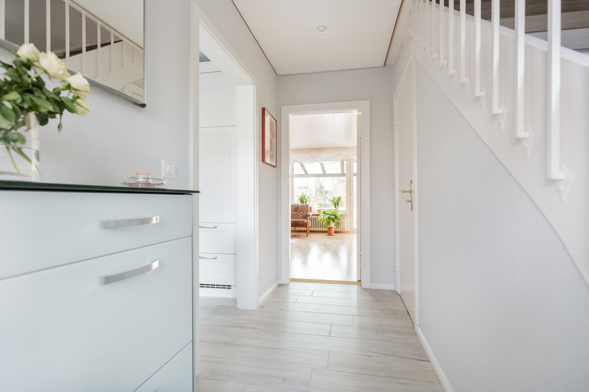 A bright, light gray hallway with a staircase, doorway, and a cabinet with flowers.