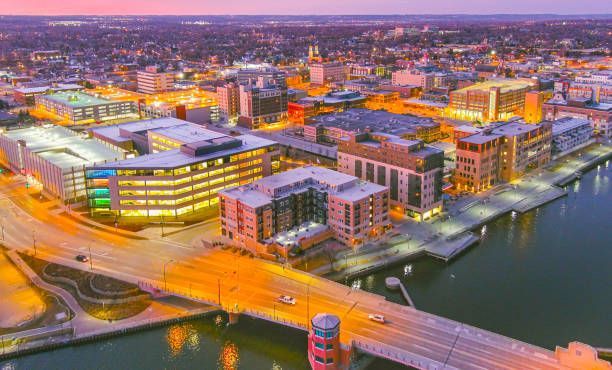 Aerial view of city buildings at dusk with lights, bridge, and water.