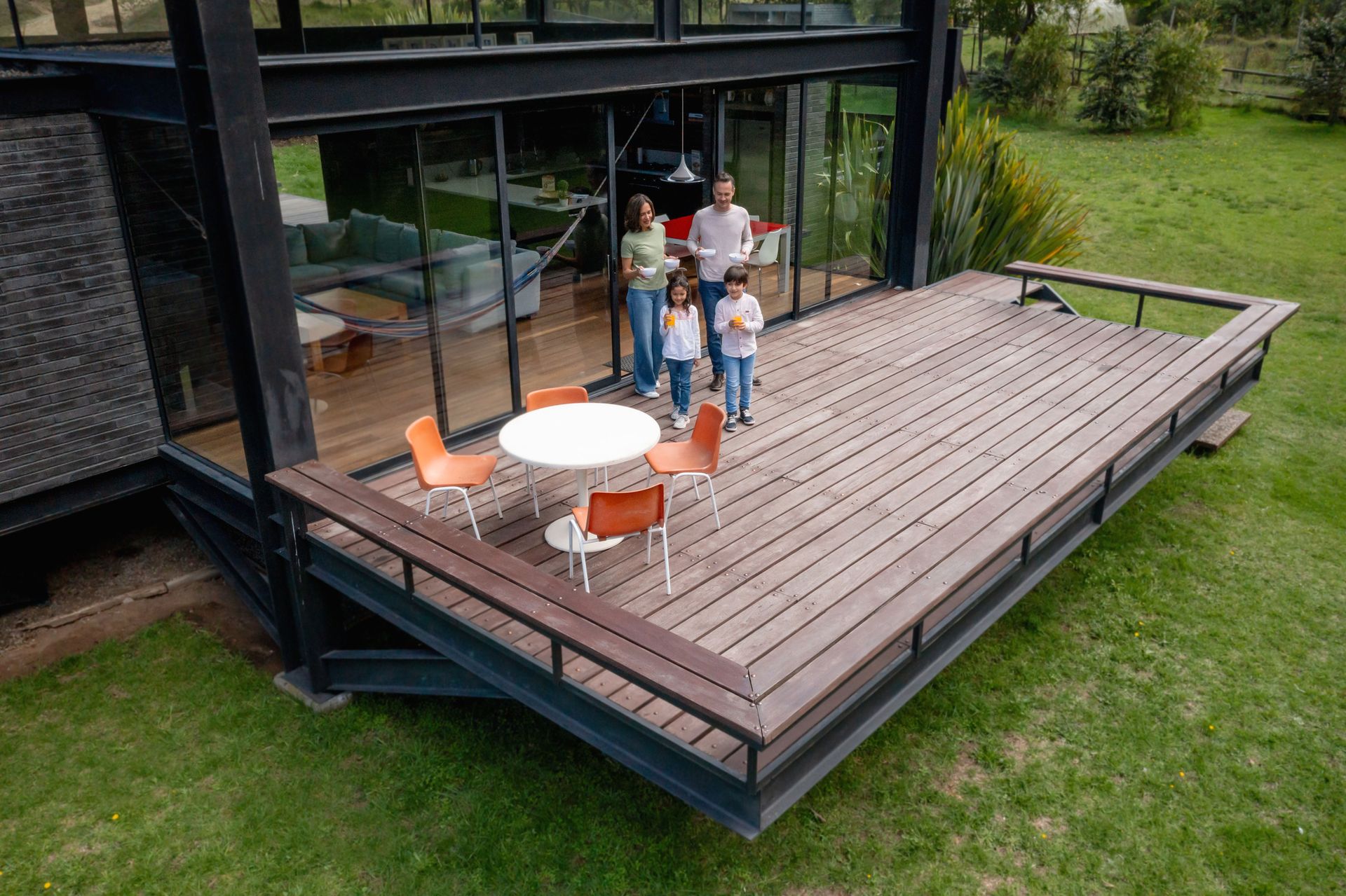 Family on a deck outside a modern house with floor-to-ceiling windows. A table and chairs are on the deck.
