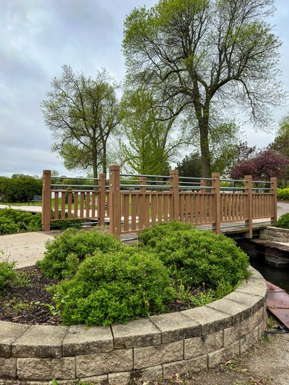 Wooden bridge over a small waterway in a park, with green shrubs in the foreground and trees in the background under a cloudy sky. Wooden bridge over a small waterway in a park, with green shrubs in the foreground and trees in the background under a cloudy sky.