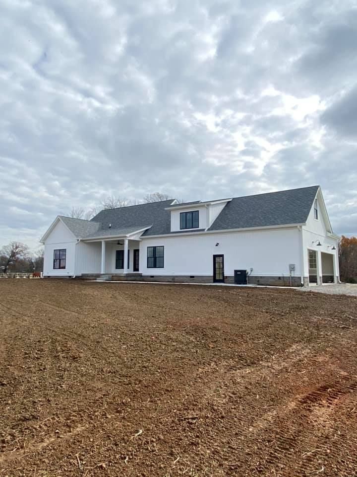 A new white modern farmhouse with a grey roof and side-entry garage sits on a plot of bare, freshly tilled brown earth.