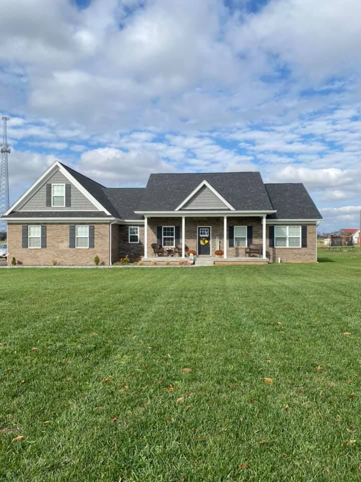 A wide, one-story brick house with a covered front porch, grey gabled roofs, and a large front lawn under a cloudy sky.