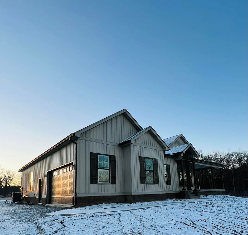 A modern, light-colored farmhouse exterior with a garage and covered porch, surrounded by snow under a clear blue sky.