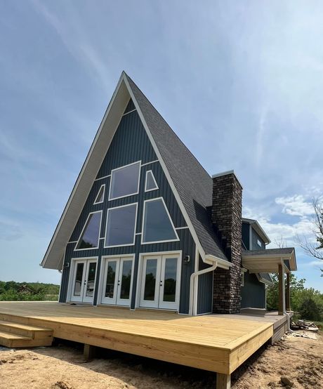 A blue A-frame cabin with large windows and a stone chimney stands on a wooden deck under a bright blue sky.