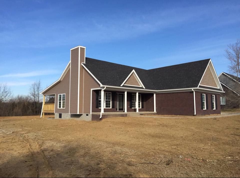 A newly constructed one-story house with dark brown brick, taupe siding, a black shingled roof, and a front porch.