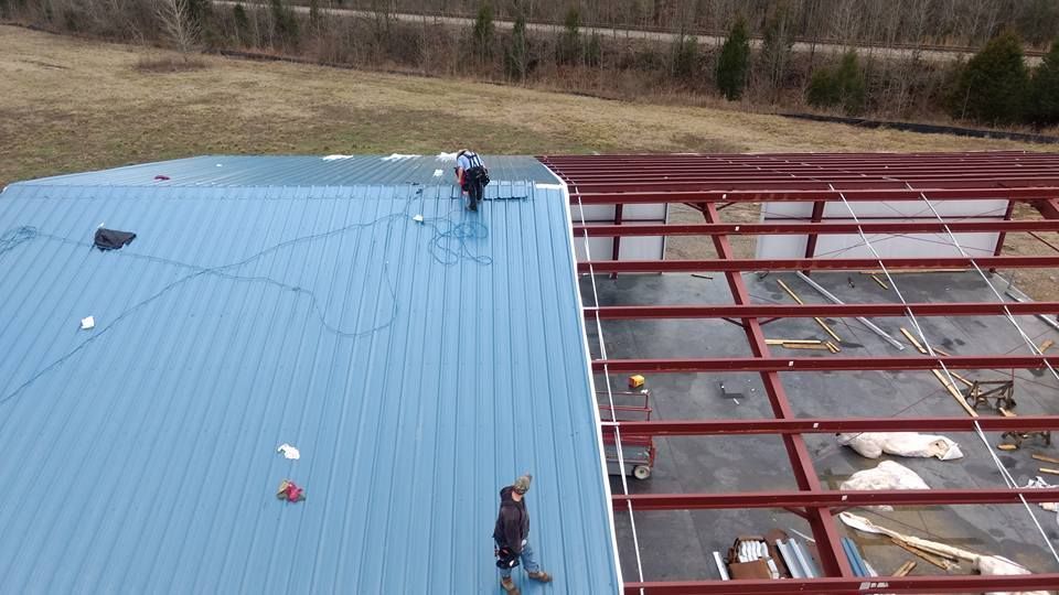 Workers install blue metal roofing sheets onto the red steel frame of a building structure under construction.