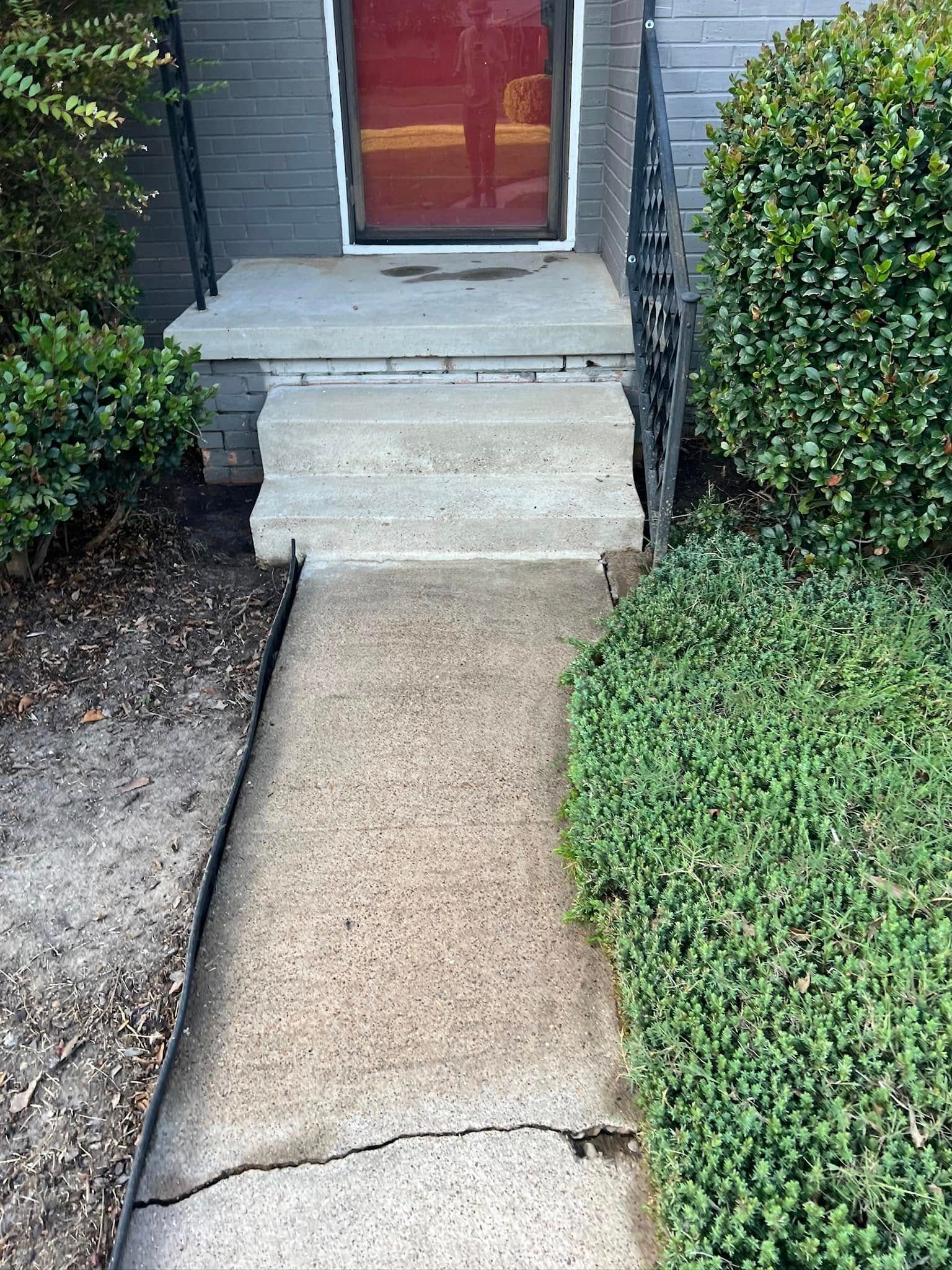 Concrete walkway leading to steps and a red door. Green bushes flank the path.