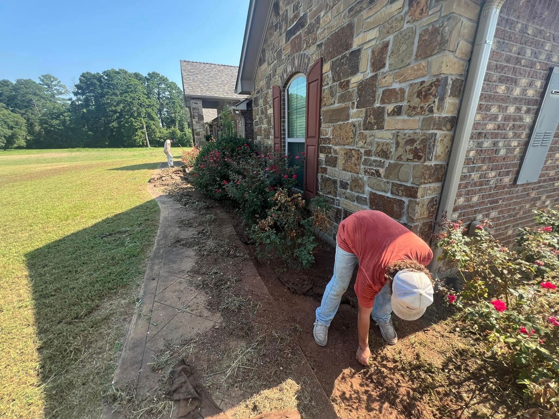 Person tending a garden bed along a brick house. They're wearing a hat and bending down near plants and flowers.