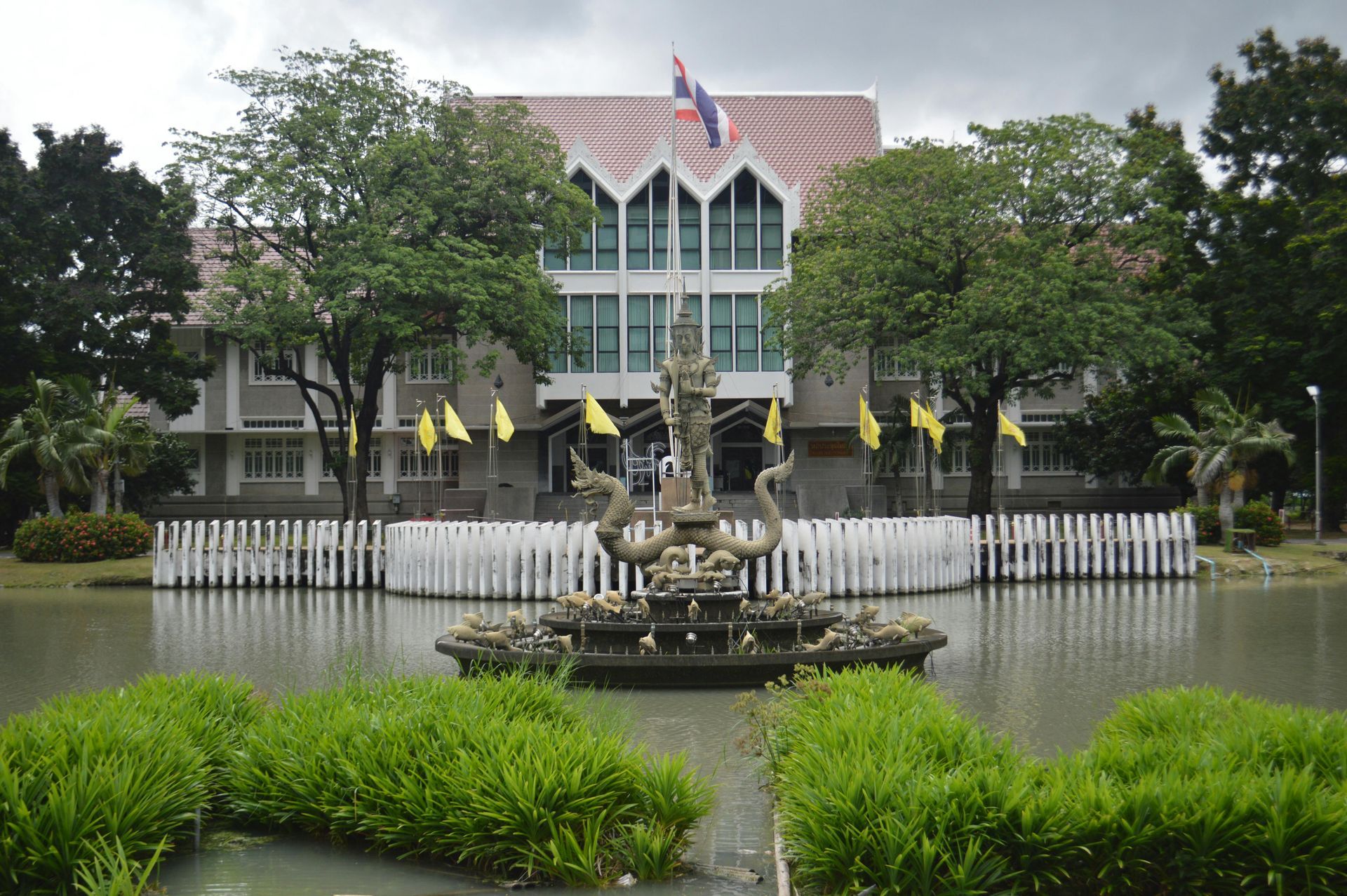 Building with Thai flag and ornate fountain in a pond, trees, overcast sky.