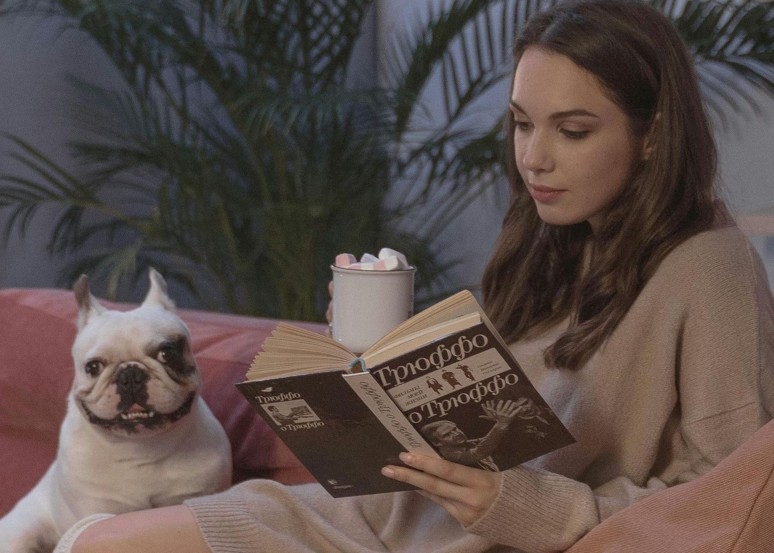 A woman is sitting on a couch reading a book next to a dog.