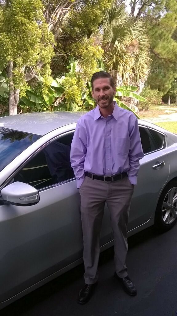 A man in a purple shirt is standing next to a silver car.