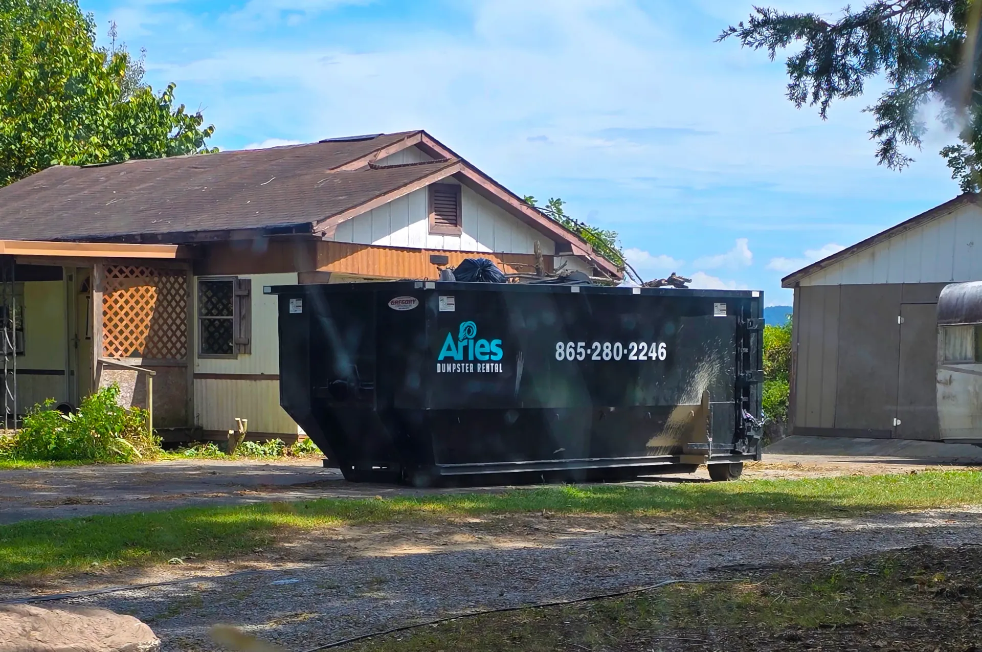 Black dumpster in front of a house; blue sky in the background, Aries Disposal logo on dumpster.