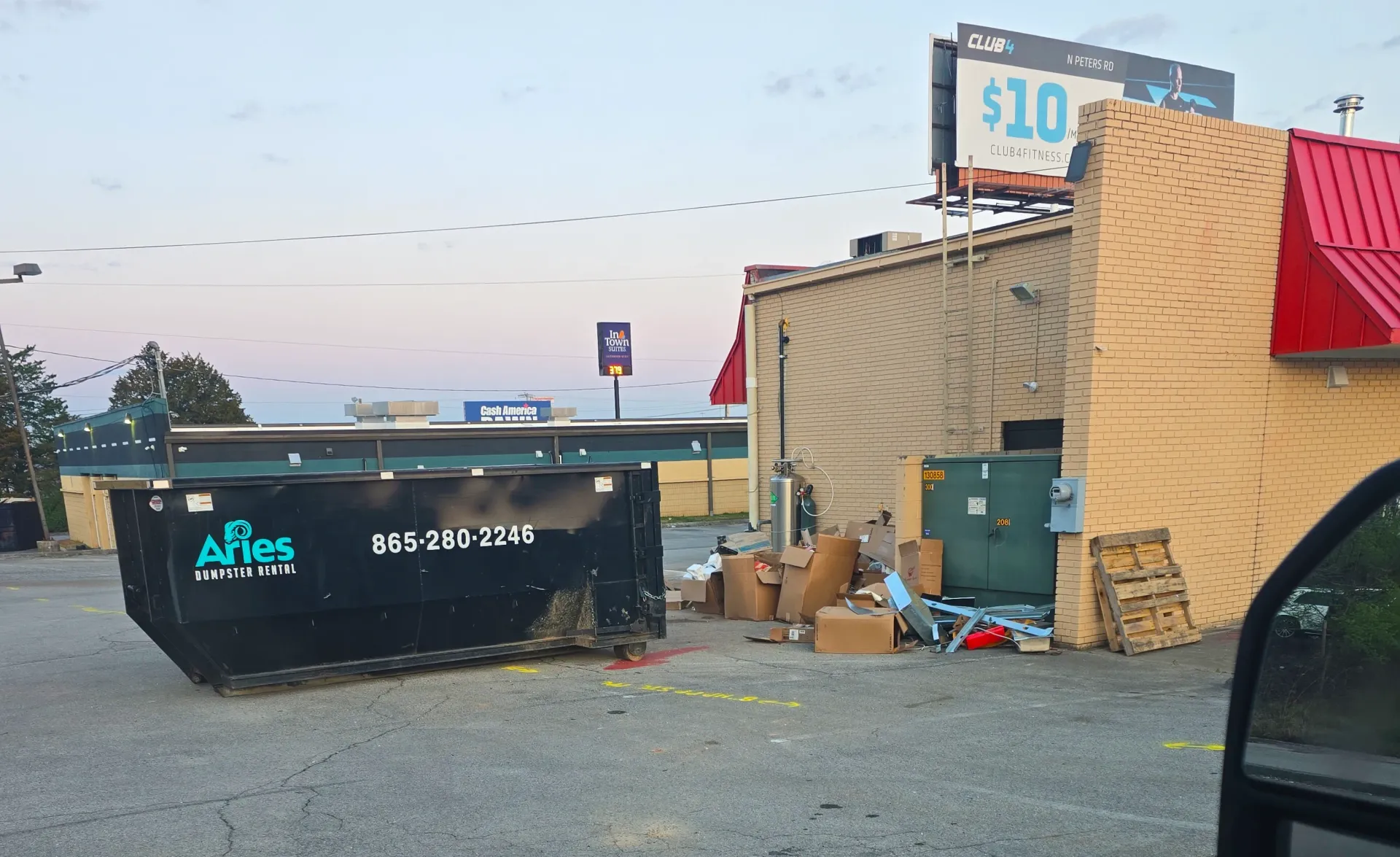 A black dumpster overflowing with trash in a parking lot next to a building. A billboard is visible.