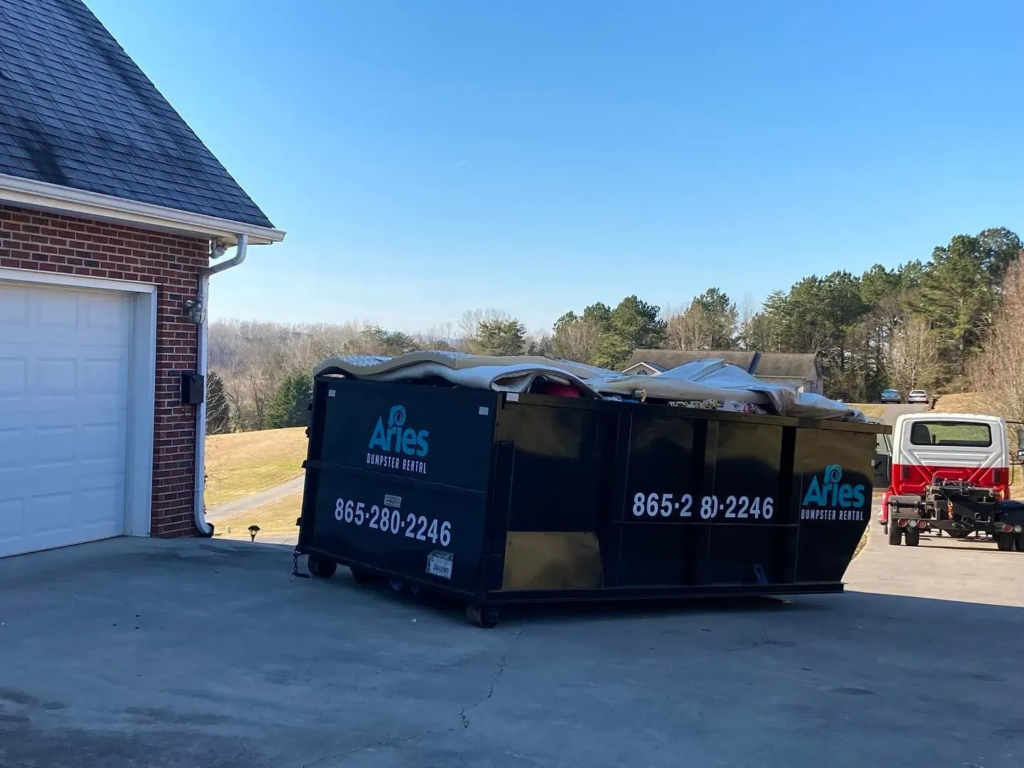 Black dumpster overflowing with debris on a driveway next to a brick garage, under a clear blue sky.