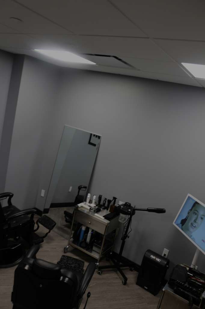 Barbershop interior with chairs, styling station, and a monitor. Gray walls and bright lighting.