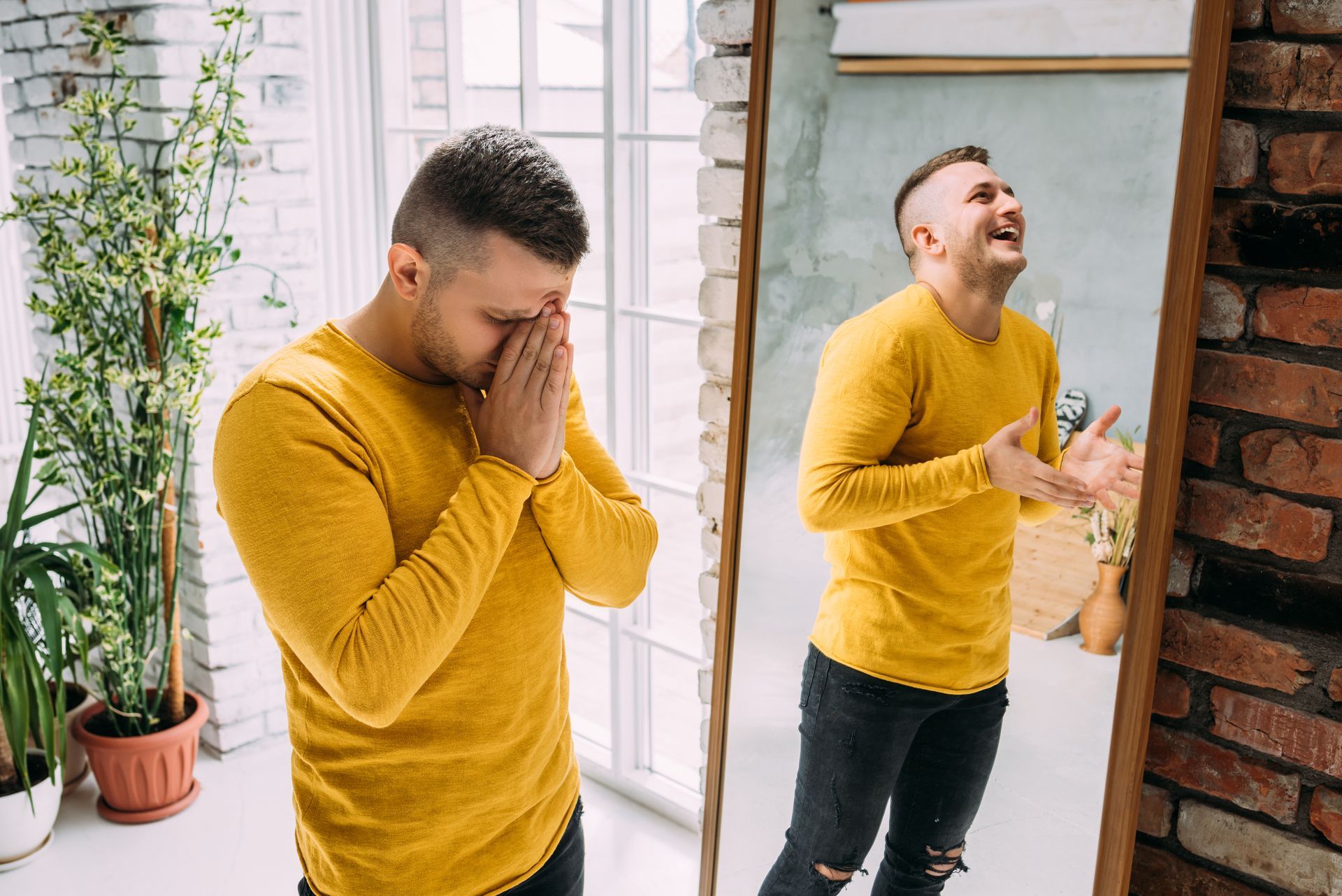 Man in yellow shirt, sad, covering face, contrasts with joyful reflection in mirror.