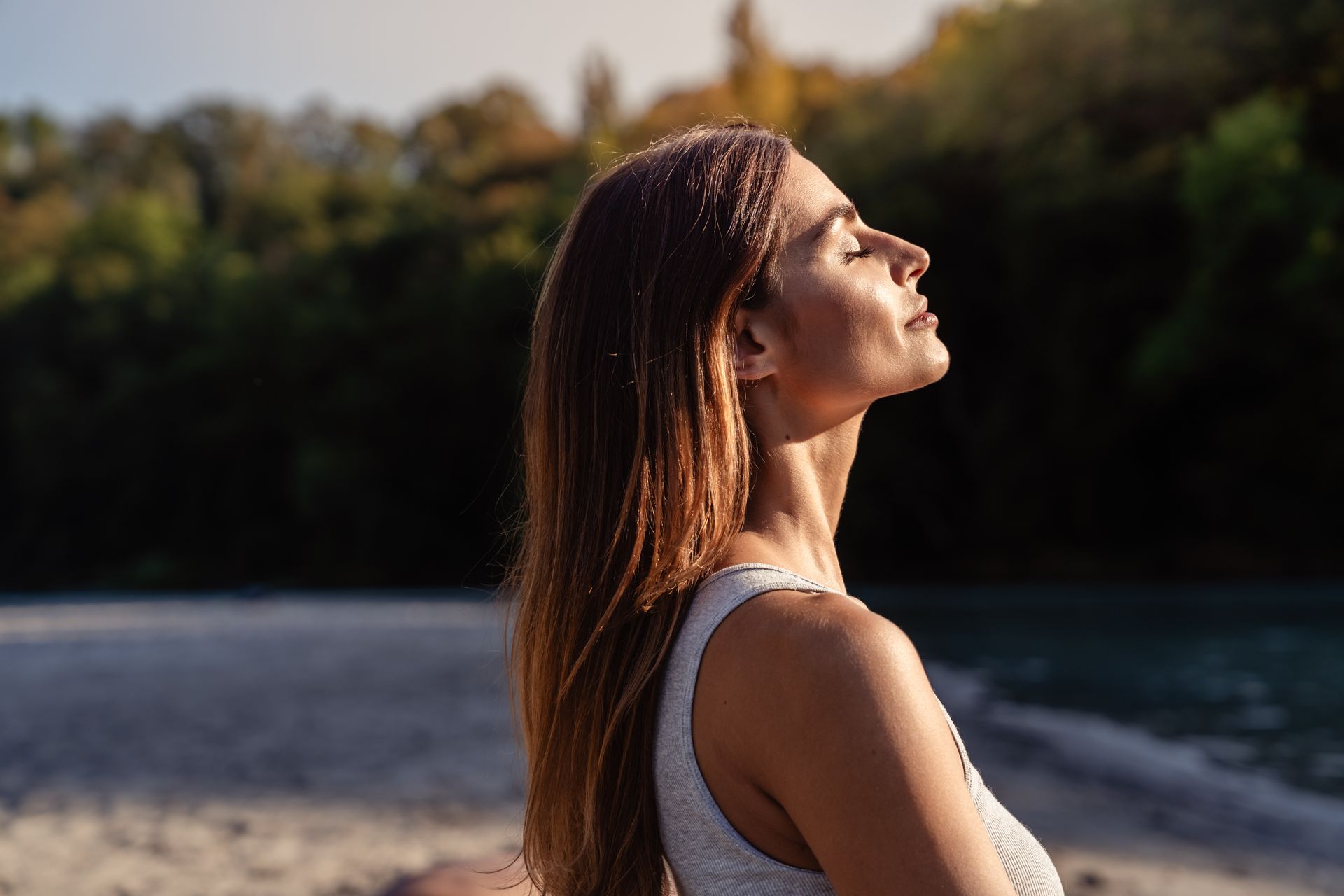 Woman with eyes closed, head tilted up, enjoying sunlight on a beach with trees in the background.