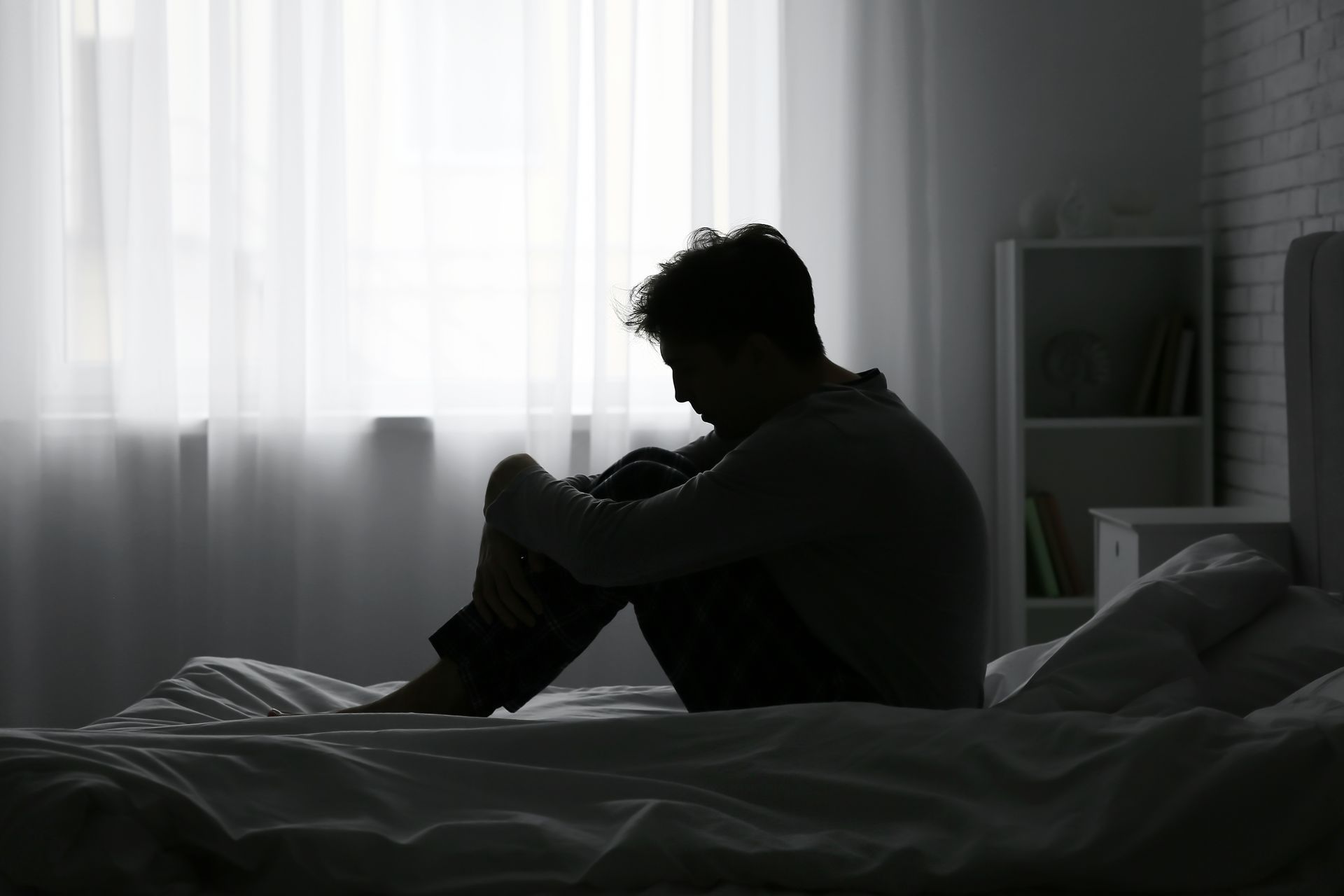 Silhouette of person sitting on a bed, head down, appearing sad, near a window.