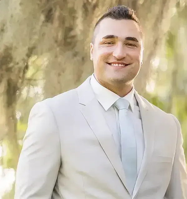 A smiling person wearing a light grey suit and white tie, posed outdoors in a blurred, sunlit, natural setting.
