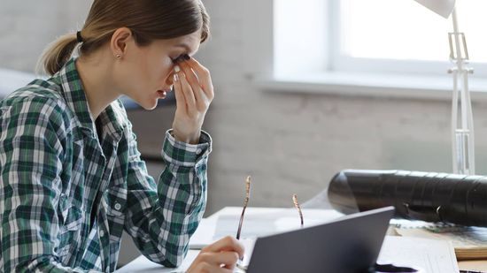 A woman is sitting at a desk with a laptop and rubbing her eyes.