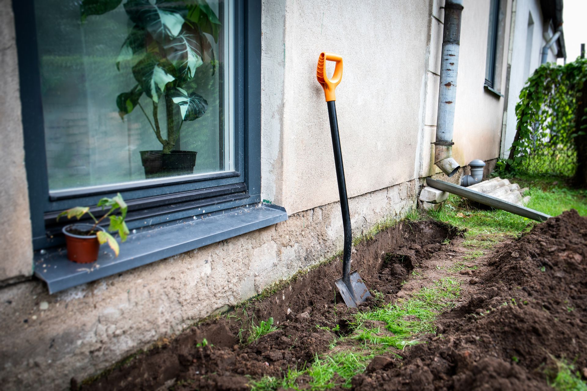 Trench dug along house foundation for basement waterproofing with shovel nearby.