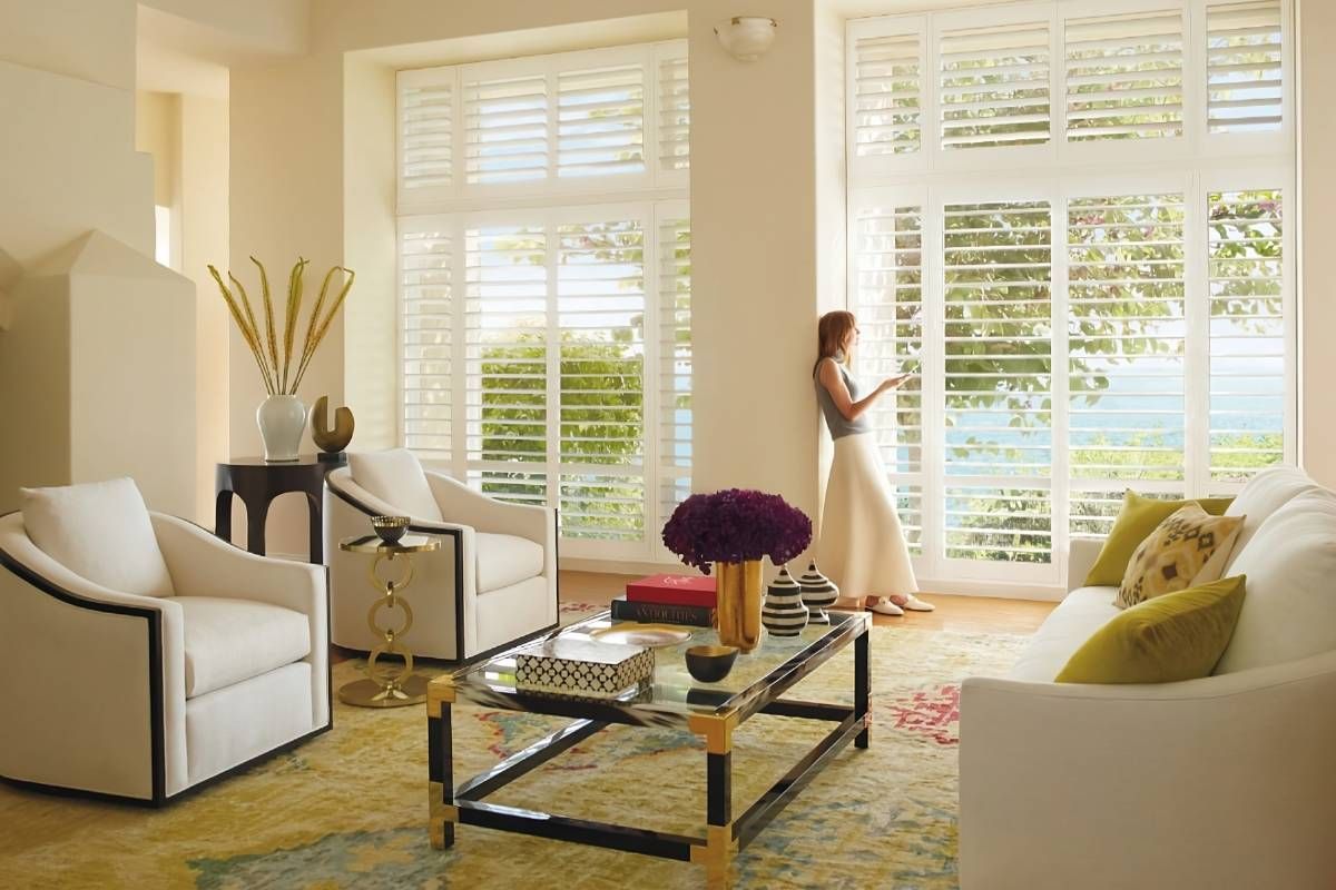 Hunter Douglas Palm Beach™ Polystain™ Vinyl Shutters as living room shades with a woman peering outside at Harmony Blinds & Shutters near San Diego, CA