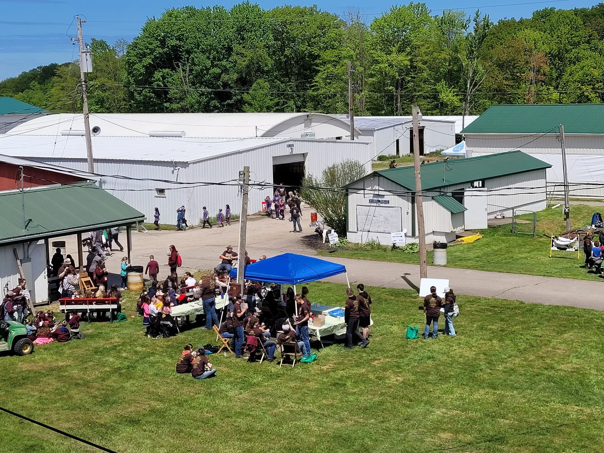 An outdoor fairground scene with people gathered near tables and a blue canopy tent amidst several large white buildings.