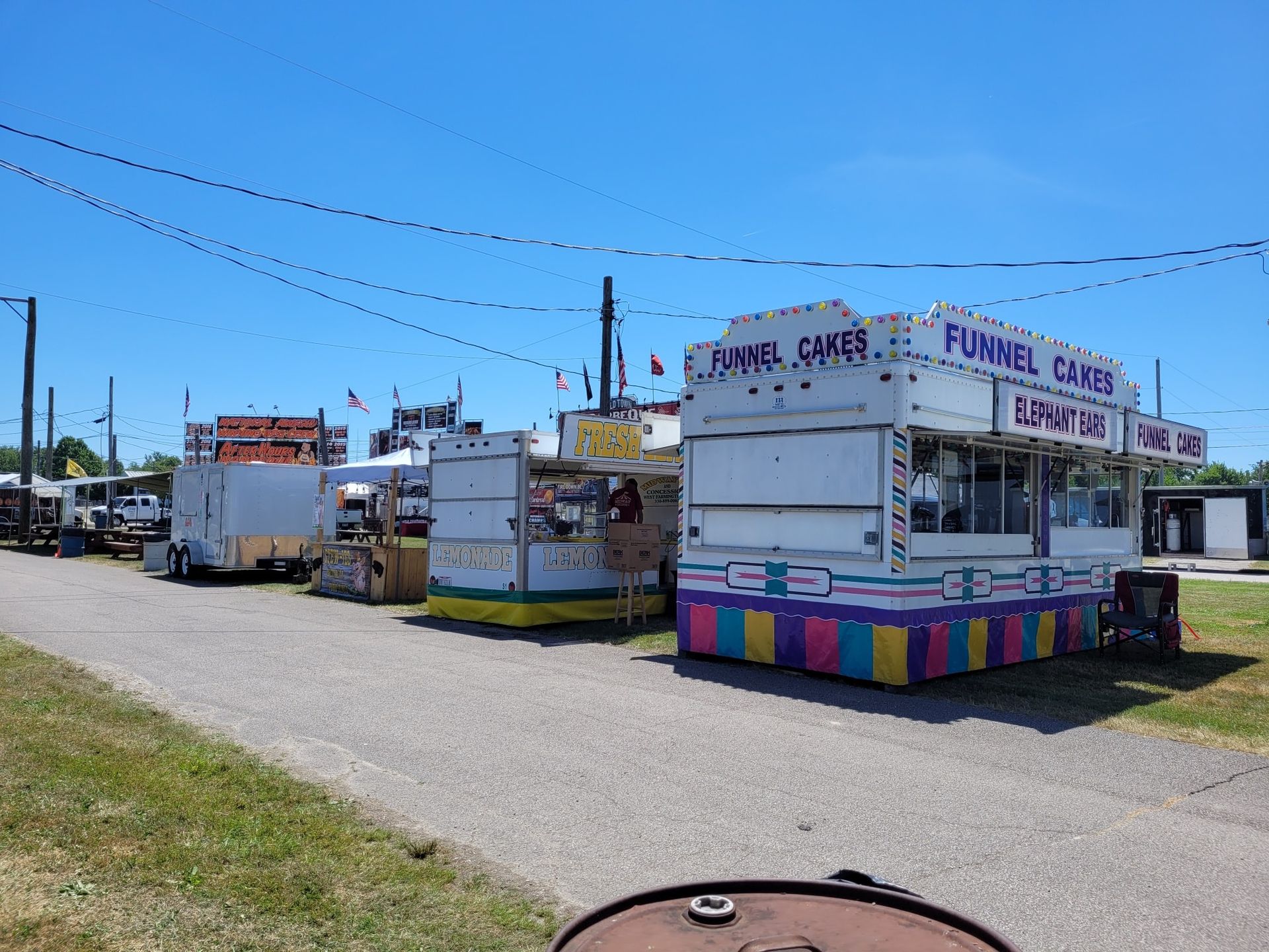 Several food and game concession trailers are lined up on a gravel path under a clear blue sky.