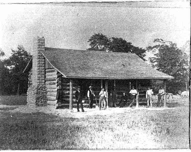 A black-and-white photo of a log cabin with a stone chimney and a striped roof, with several people standing on the porch.