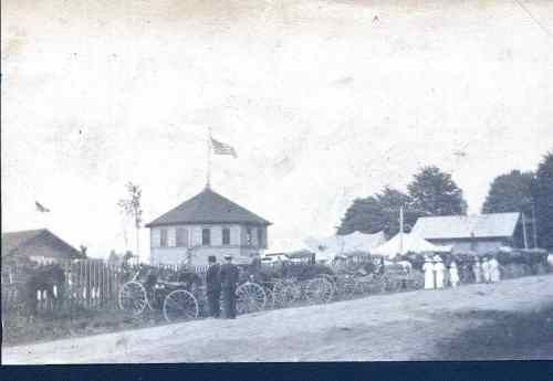 A historical black-and-white photo of a multi-sided pavilion with a flag, horse-drawn carriages, and people standing nearby.