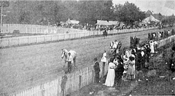 Historical scene of a dirt racetrack with a person leading a horse, lined by a wooden fence and spectators.