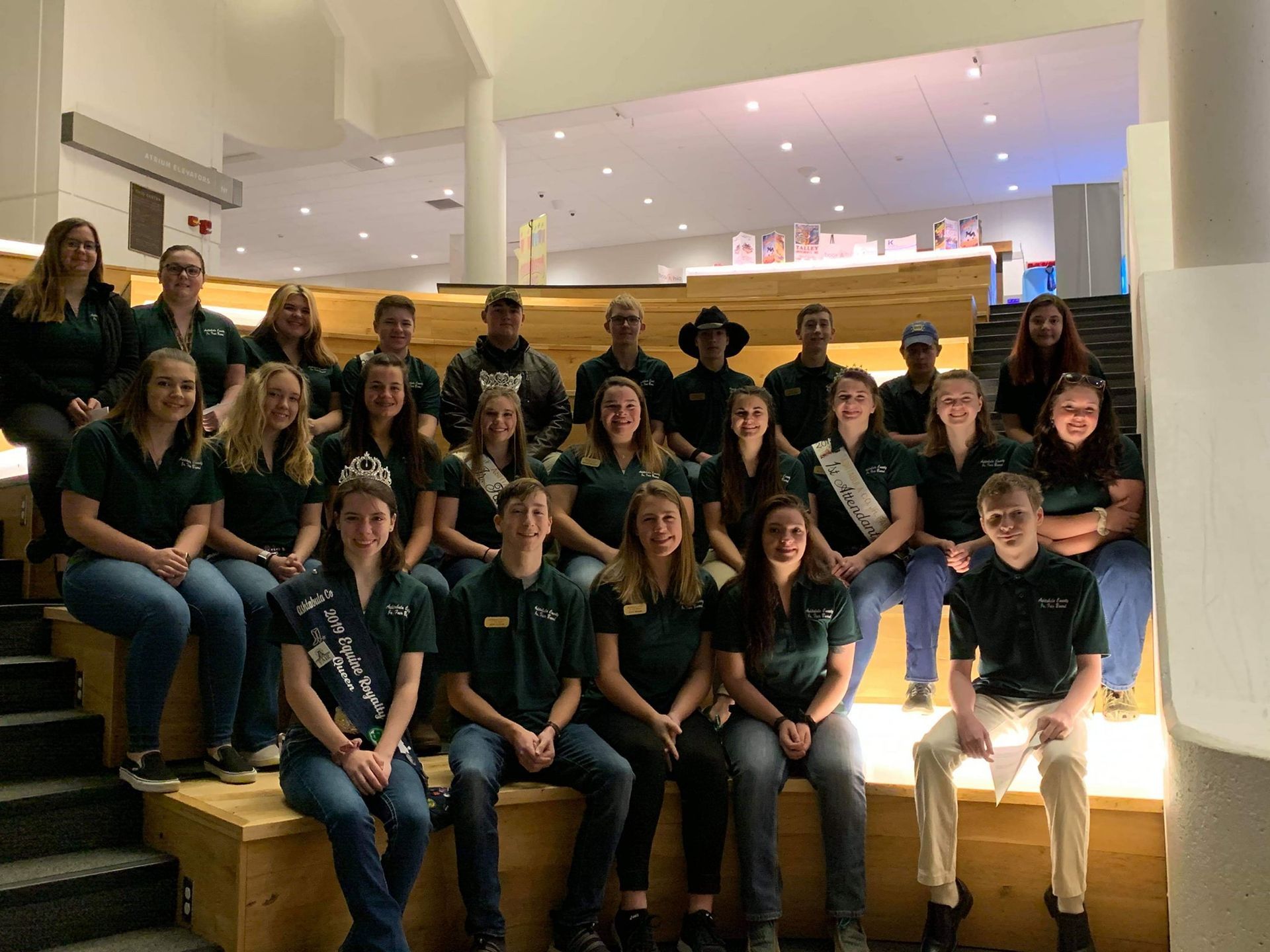 A group in matching dark green polos poses on tiered wooden seating in a modern, brightly lit interior space.