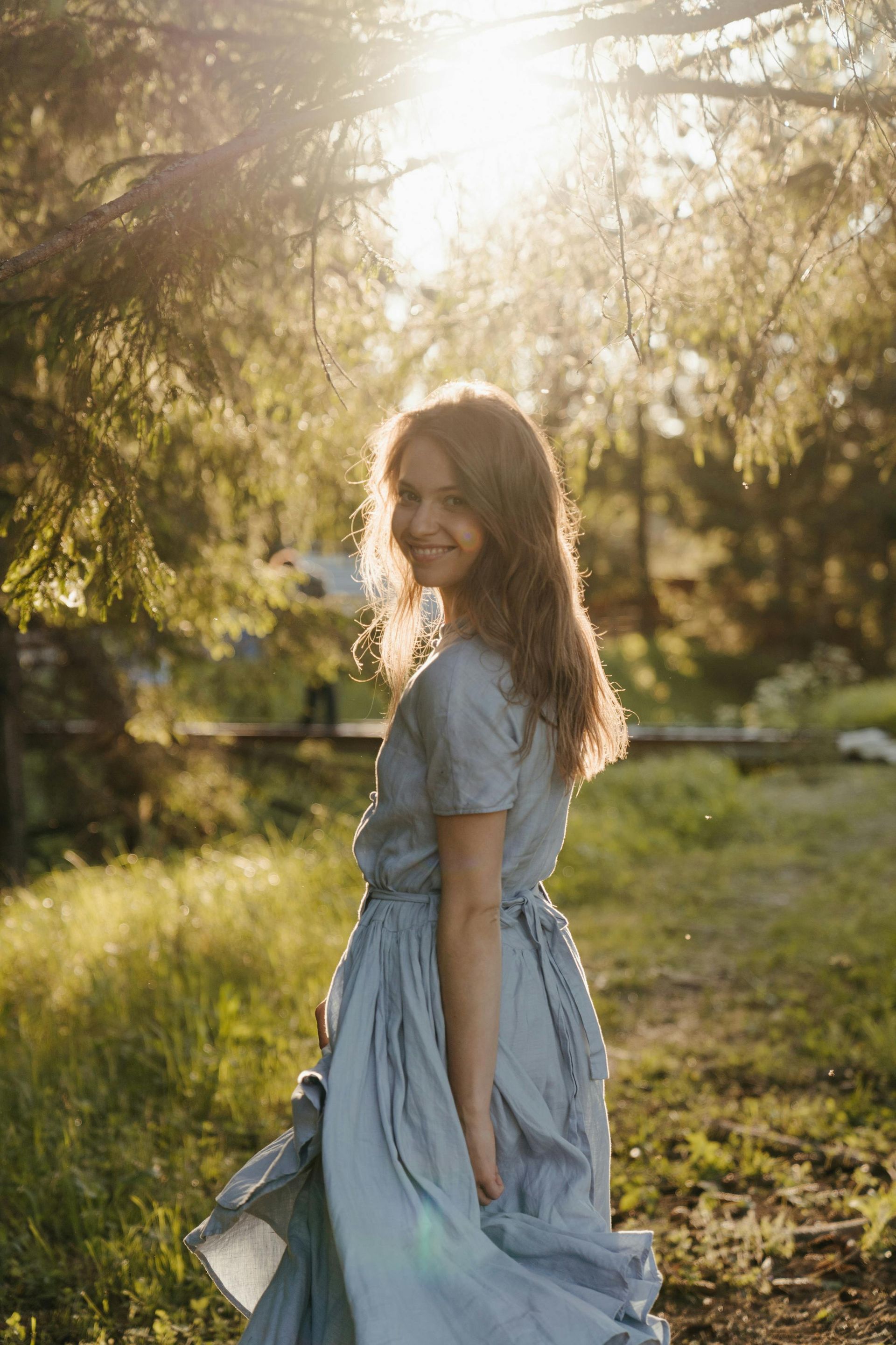 A person in a light blue dress walking through a sunlit field, looking back over their shoulder with a soft smile.