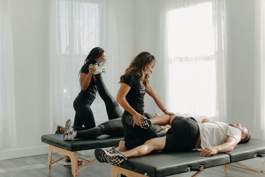 Two therapists stretching and examining a patient's leg on a massage table in a bright room. A third therapist looks on.