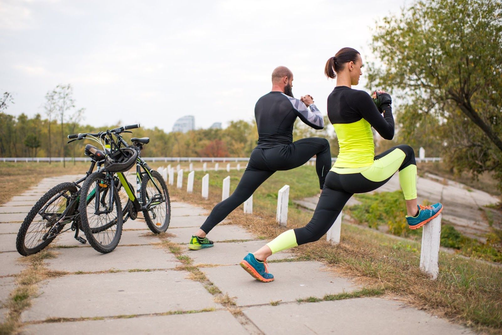 Two cyclists are doing warm-up stretches before a ride in the park.
