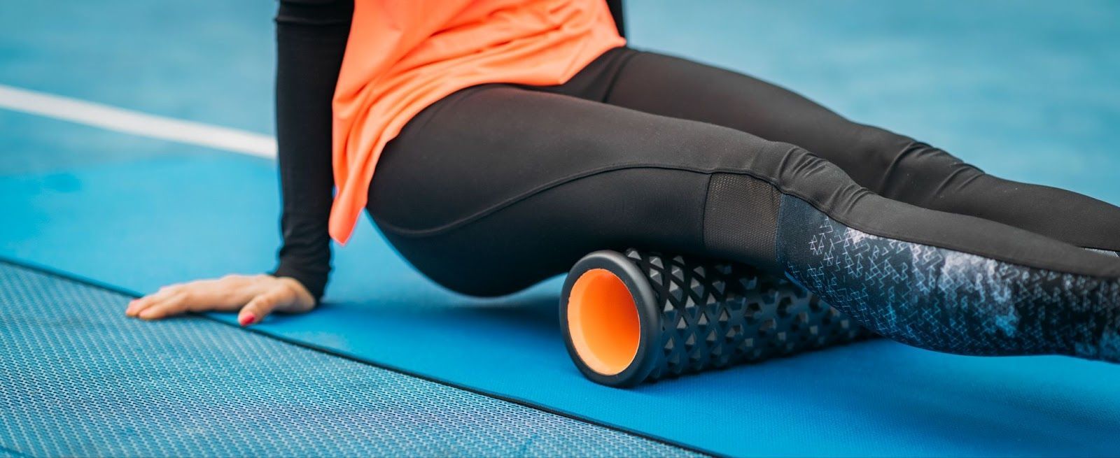 An unidentified female athlete in black and orange gear uses a foam roller for stretching exercises.
