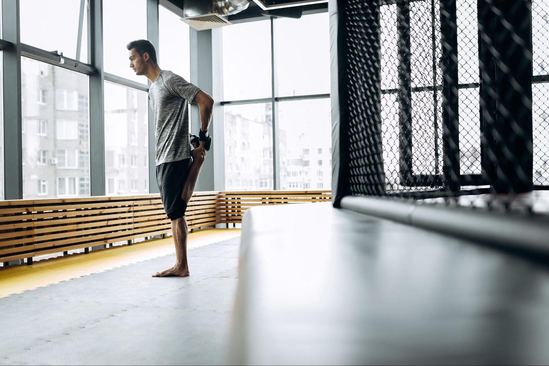 A man stretches his left leg as he looks out the window at an urban cityscape.