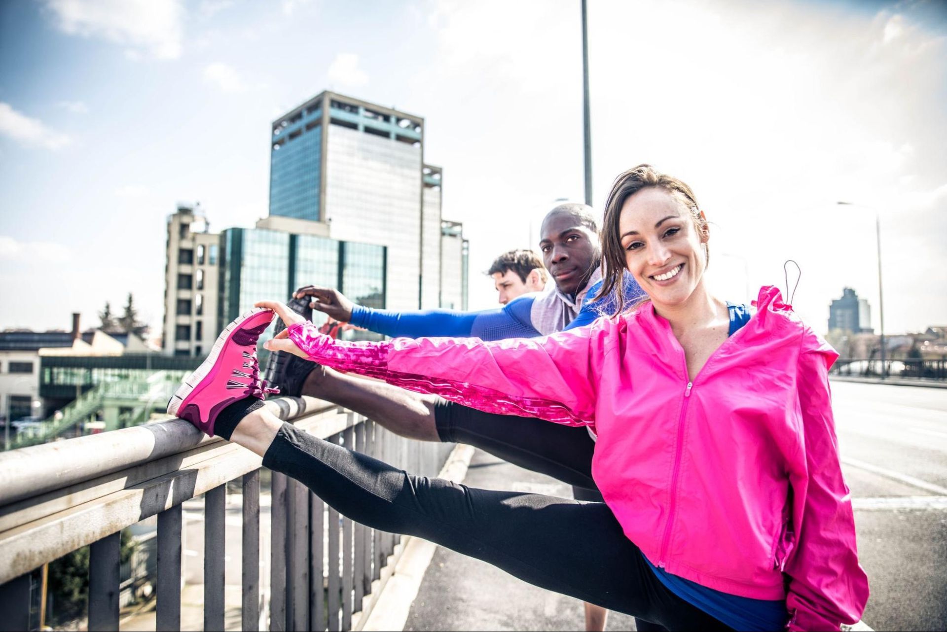 A woman and two men stretching their leg on the side of a bridge with a street and buildings in the background. 