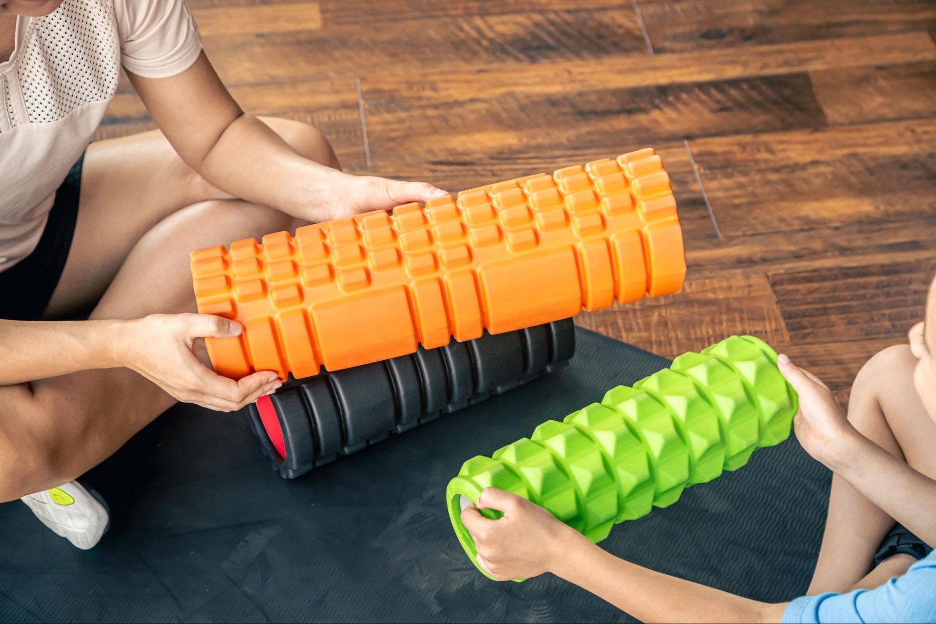 A woman and a little girl look at three different foam rollers together while sitting on the floor.