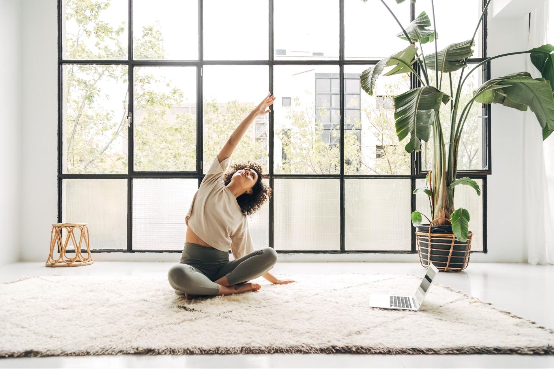 A woman stretches in her home, in front of a large window.