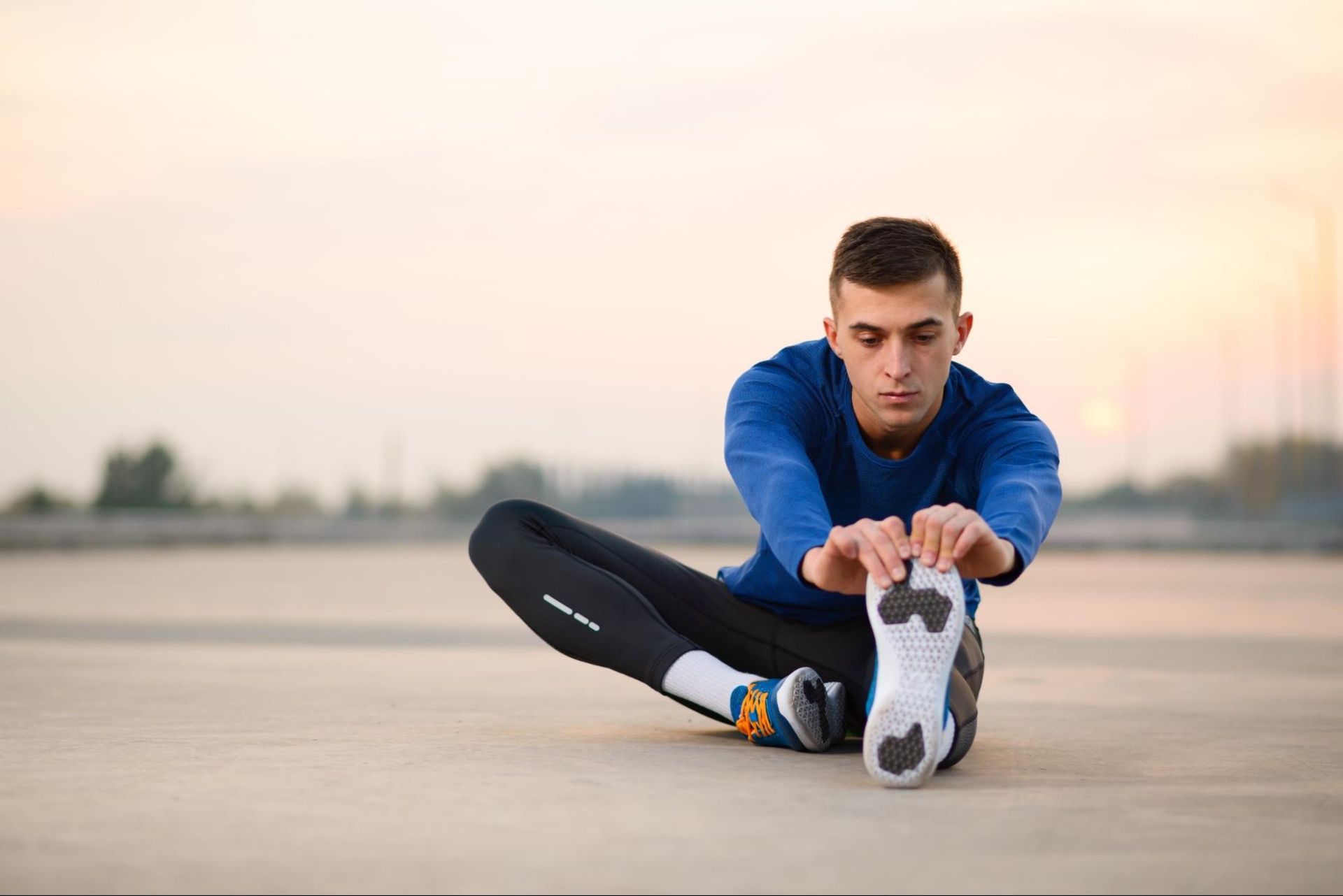 A young man in running clothes stretches his legs while sitting on the ground. 