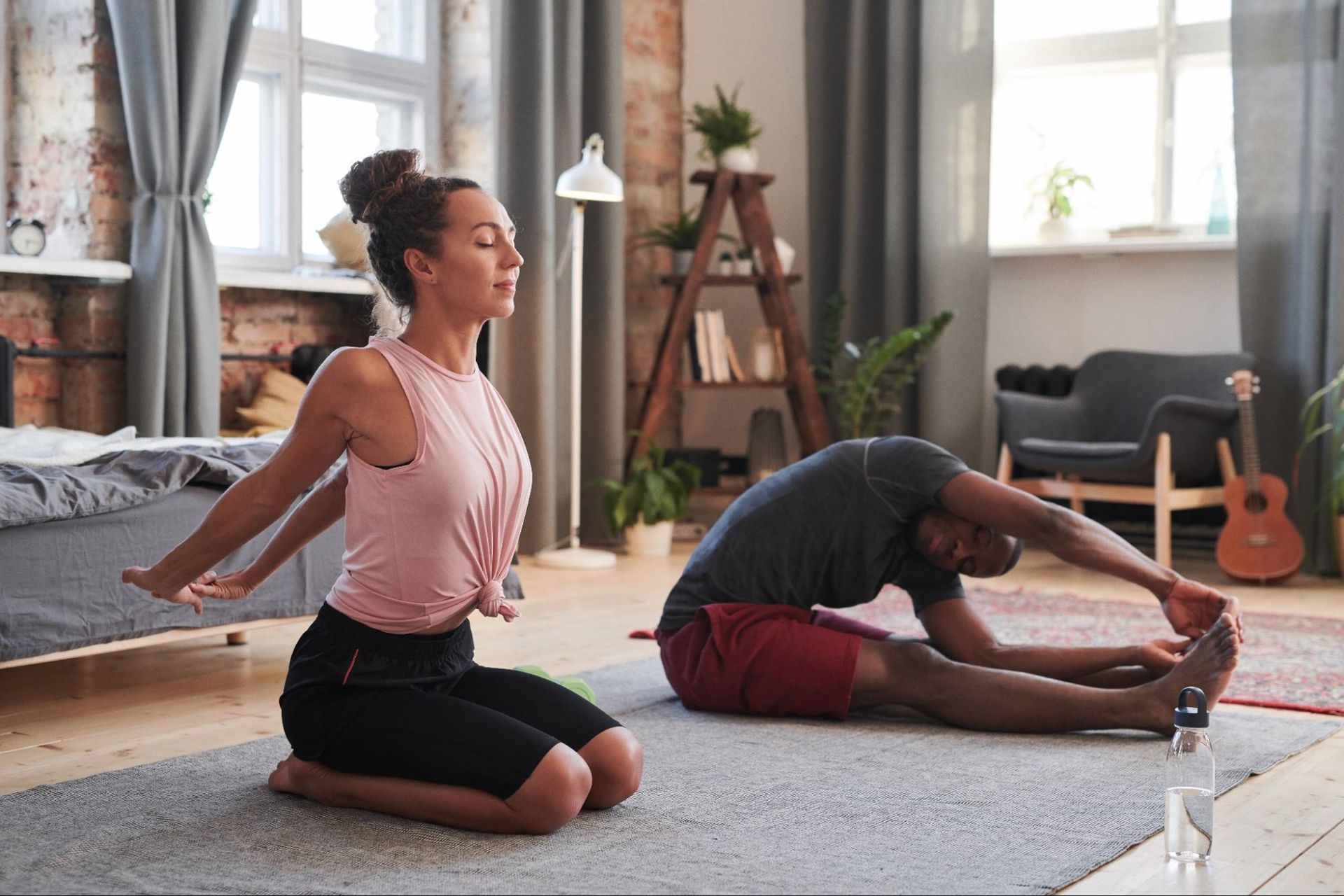 A couple stretches at home together on a rug. A woman stretches her arms behind her back while a man stretches his legs.